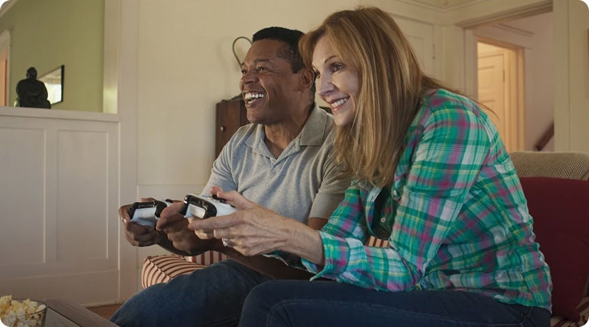 Two people sitting on a couch, smiling and playing a video game together with controllers 