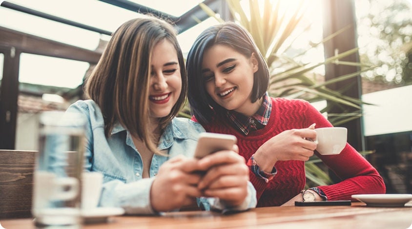 Two friends sitting at a café table, smiling as they look at a smartphone together while drinking coffee. 