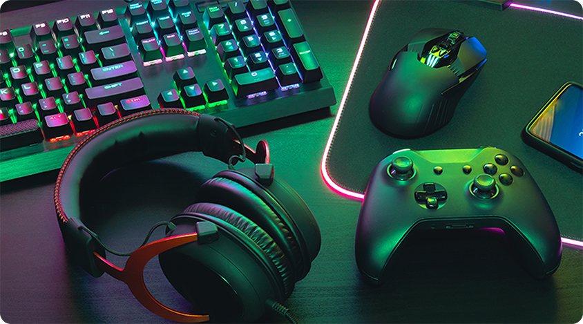 Gaming setup on a desk featuring a keyboard, mouse, controller, headset, and phone illuminated by RGB lighting