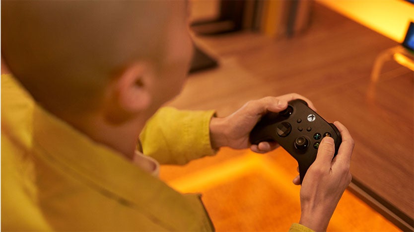 A person wearing a yellow jacket is holding a black gaming controller while playing a video game. The image is taken from behind and slightly above, showing their hands gripping the controller.