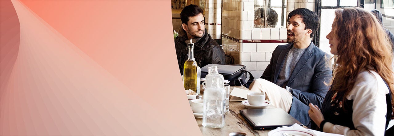 A group of three people sits around a table in a cozy cafe with tiled walls. One person in a blazer is speaking animatedly to the group, while another person, dressed in a dark jacket, listens attentively. A third person with long, wavy hair sits to the right, also listening.