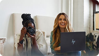 Two people, one holding a camera and pointing it forward, the second person is holding a laptop and smiling.