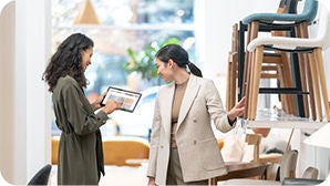 Two people in a furniture store, one holding a tablet and interacting with the other who is gesturing towards products on display.