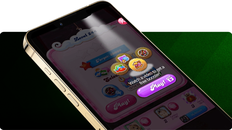 Smartphone angled on its side displaying a candy-themed game screen with a prompt that says “Watch a video to get a free booster,” featuring colorful game icons and a purple “Play!” button, set against a green striped background.