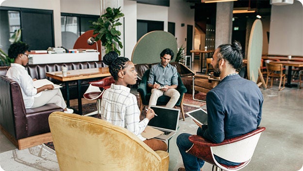 Four people sitting in a room reading, writing using a laptop and a tablet.