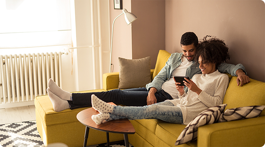 Two people relaxing on a couch, smiling and looking at a smartphone together in a cozy living room