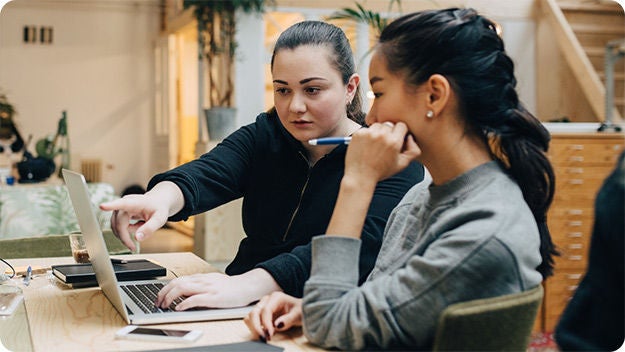 Two people sitting indoors at a desk working on a laptop computer.