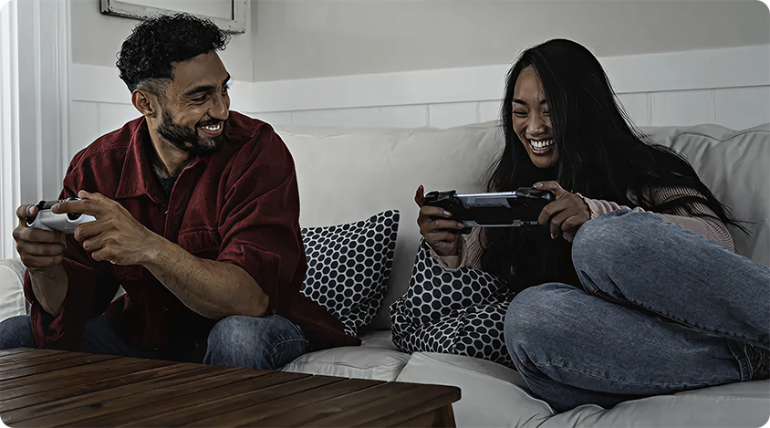 Two people smiling and playing games together on handheld gaming devices while sitting on a couch. 