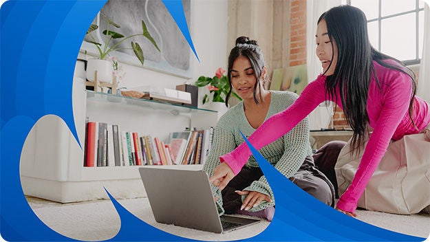 Two people are sitting on the floor in a living room, looking at a laptop placed in front of them.