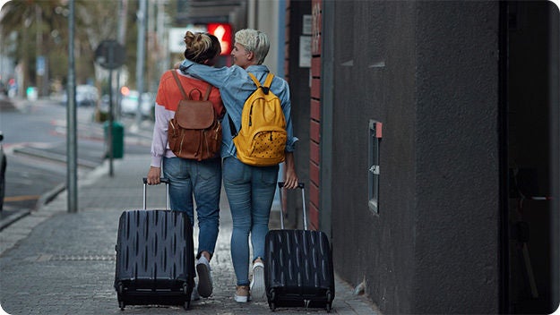 Two people are walking down a city street, each pulling a suitcase. They both have backpacks and are dressed casually in jeans and jackets. One person has an arm around the other.