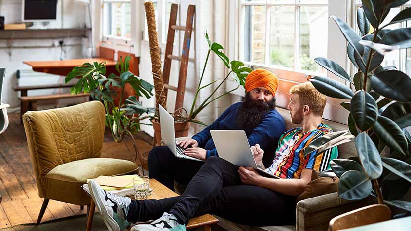 Two men in an apartment sit on a couch and work on laptop computers.