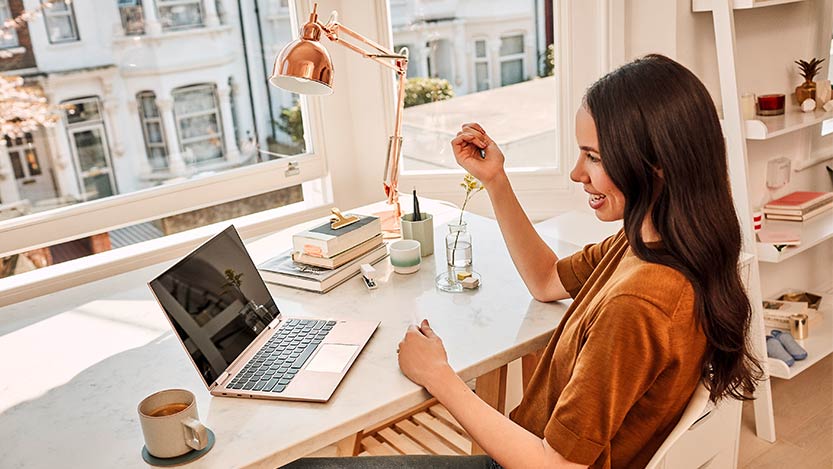 A woman sitting at a desk near a window works on her laptop computer.