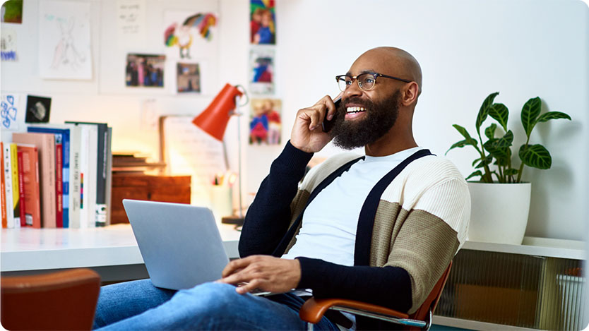 A man smiling and working at a home office while talking to the phone.