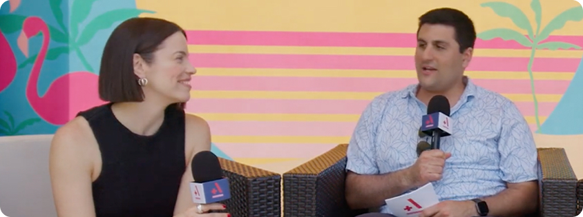 Two people sitting together and speaking into microphones during an on-camera interview at an event.