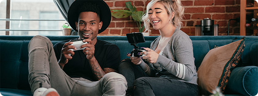 Two people sitting on a couch playing games together, one on a console controller and the other on a mobile device with a clip attachment.