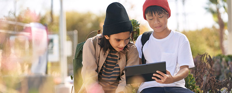 Two children sitting outside look at a tablet.