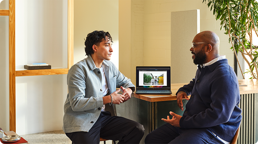 Two professionals sitting and talking at a table in a modern office, with a laptop open between them showing a digital interface.