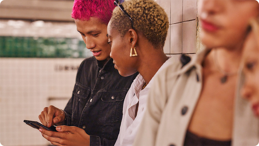 Three people standing in a subway station, one person is holding and looking at a smartphone. The background shows tiled walls and part of the subway platform.