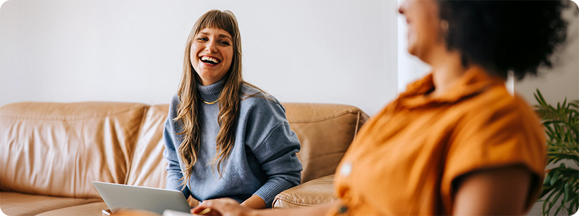Photograph of two women sitting on a brown leather couch, engaged in a cheerful conversation. One woman wears a blue sweater and holds a laptop, while the other wears an orange top.