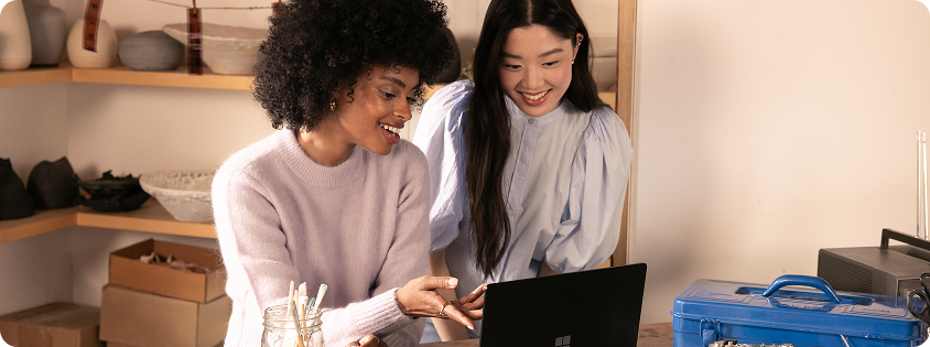 Photograph of two women sitting at a wooden table, looking at a laptop and smiling, suggesting collaboration or shared interest.