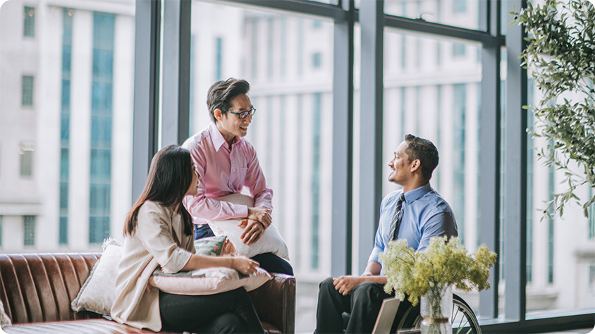 Photograph showing three professionals engaged in a discussion in a modern office with large windows and cityscape view.