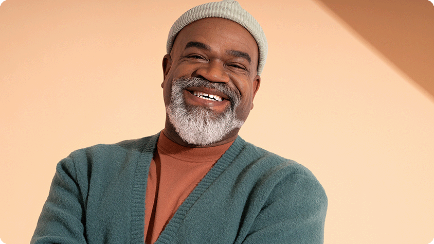 Photograph of a person wearing a teal cardigan, rust-colored shirt, and light gray beanie standing against a peach-colored background with diagonal shadow.